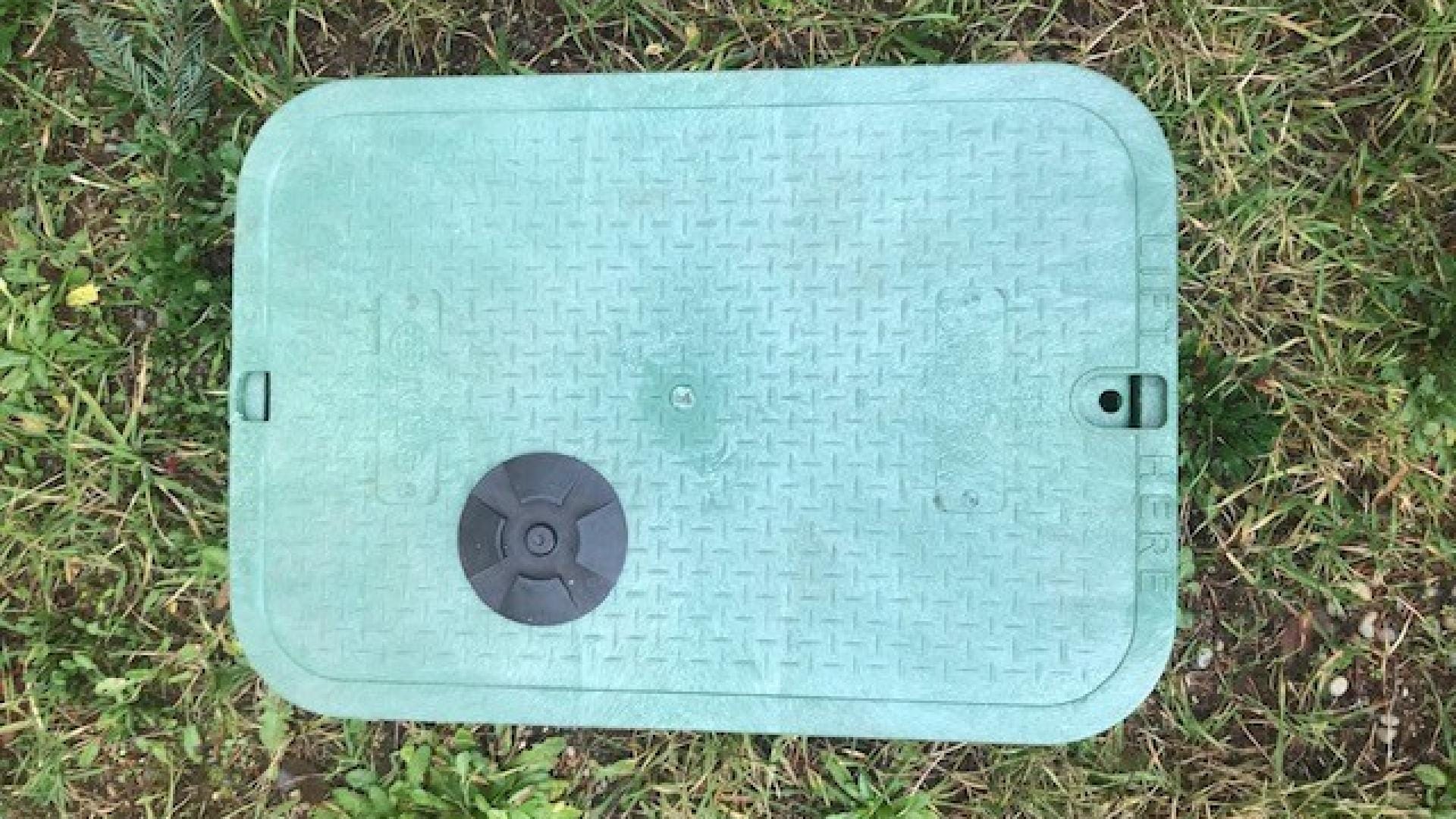 Greenish water meter box on ground. The water meter box is surrounded by grass.